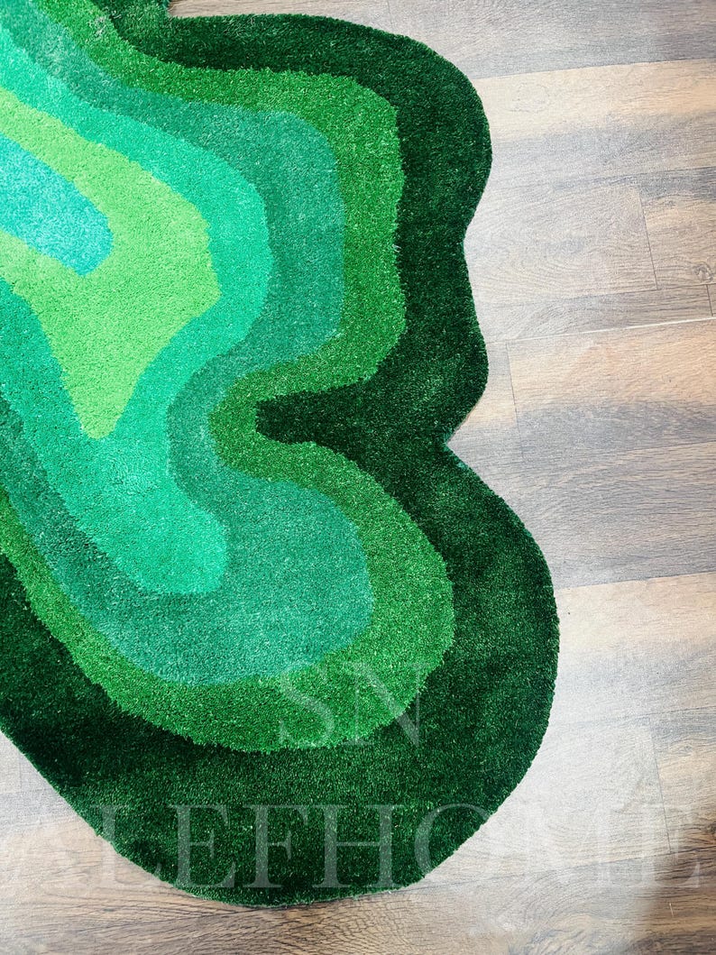 May include: A green, abstract-shaped rug with varying shades of green, from light to dark. The rug has a soft, textured surface and is placed on a wooden floor. The design features layered, organic shapes.