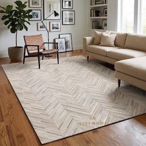 May include: A rectangular area rug featuring a light-colored, multi-tonal herringbone pattern made of thin, linear strips. The rug displays a neutral palette of cream, beige, and soft gray tones, covering a large portion of the wooden floor.