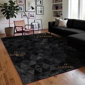 Peut inclure: Un grand tapis rectangulaire présentant un motif géométrique de triangles noir foncé sur toute sa surface. Le tapis est posé sur un parquet dans une pièce avec un canapé noir, une chaise en bois et une plante. Le texte SN ALEF HOME est visible.