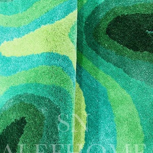 May include: A close-up of a plush rug with a swirling pattern in shades of green and teal. The rug's design features concentric bands of varying shades, creating a textured, layered effect. The rug has a soft, inviting appearance.