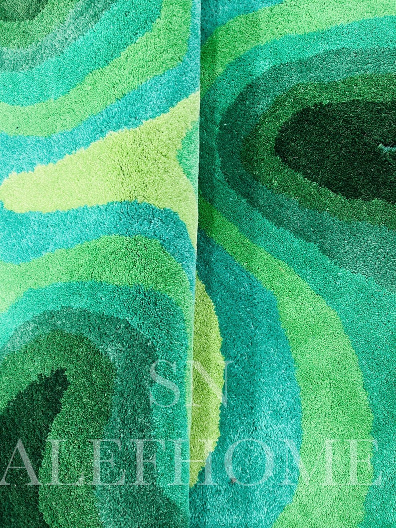 May include: A close-up of a plush rug with a swirling pattern in shades of green and teal. The rug's design features concentric bands of varying shades, creating a textured, layered effect. The rug has a soft, inviting appearance.