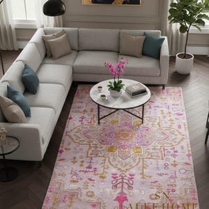 Puede incluir: Una alfombra rosa claro con un patrón simétrico detallado en dorado y fucsia. La alfombra está colocada en una sala de estar con un sofá seccional gris, una mesa de centro redonda y una planta en maceta. La alfombra tiene el texto "SN DALEE HOME".