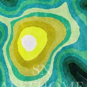 May include: A colorful rug with a layered design in shades of green, yellow, and blue. The rug features a circular pattern with a bright yellow center, surrounded by concentric rings of varying green and blue hues. The text "SN ALL HOME" is visible.