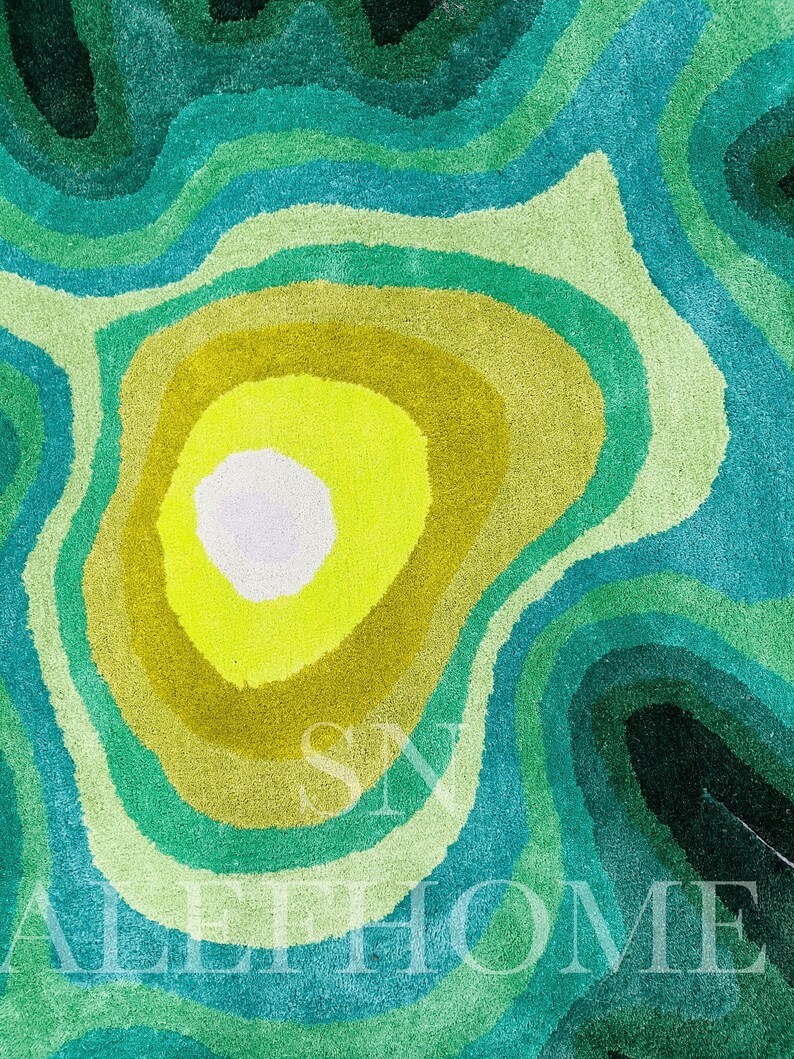 May include: A colorful rug with a layered design in shades of green, yellow, and blue. The rug features a circular pattern with a bright yellow center, surrounded by concentric rings of varying green and blue hues. The text "SN ALL HOME" is visible.