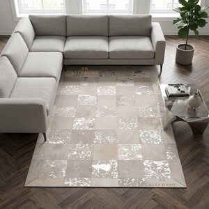 Op de afbeelding: Een rechthoekig vloerkleed met een dambordpatroon van afwisselende lichtbeige en metallic zilveren koeienhuidstukken. Het kleed bevat de tekst SN ALEF HOME in twee hoeken en ligt op een donkere houten vloer in een woonkamer.