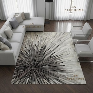 Puede incluir: Una alfombra rectangular con un patrón de rayos de sol compuesto por estrechas tiras de piel cosidas en tonos negro, gris y blanco. La alfombra está colocada en una sala de estar con un sofá seccional gris claro y dos sillones.