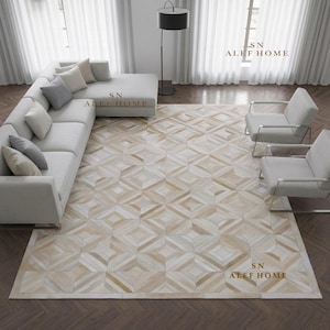May include: A large area rug featuring a geometric pattern of interlocking diamonds and squares in shades of cream, beige, and light brown. The rug is positioned in a living room with a light gray sectional sofa and two matching armchairs.
