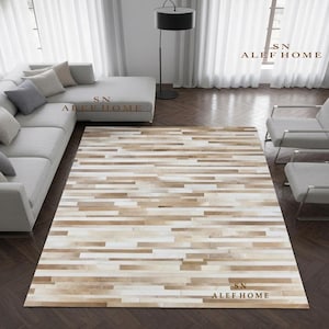 May include: A rectangular area rug featuring a horizontal striped pattern composed of alternating strips of white, beige, and light brown leather. The rug is positioned on a dark wood floor in a room with a gray sectional sofa and two armchairs.
