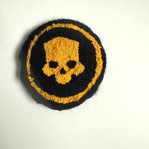 May include: A round, black and yellow patch featuring a skull design. The skull is yellow and centered within a black circle, which is surrounded by a yellow border. The patch is likely made of fabric or yarn.