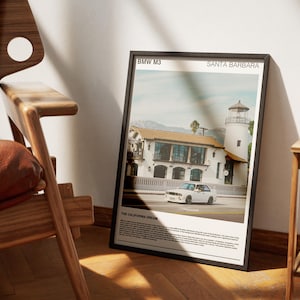 Könnte beinhalten: Gerahmter Kunstdruck mit einem weißen BMW M3 in Santa Barbara, Kalifornien. Das Bild zeigt ein Gebäude mit Leuchtturm und den Text "THE CALIFORNIA DREAM". Der Druck wird neben einem Holzstuhl angezeigt.