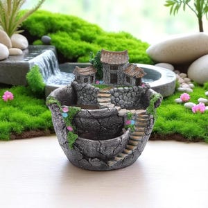 Op de afbeelding: Een grijze, steenachtige plantenbak met een miniatuur dorpsscène. De plantenbak heeft een gelaagd ontwerp met trappen, een klein huis en decoratieve bloemen. De achtergrond omvat een miniatuur waterval en weelderig groen.