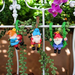 Puede incluir: Tres coloridos gnomos de jardín colgando de una valla metálica blanca. Un gnomo sostiene una fresa roja, otro está suspendido de un poste de madera y el tercero está en un columpio verde. Los gnomos tienen barbas blancas y usan sombreros.