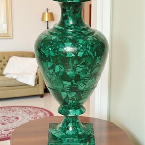 Op de afbeelding: Een grote, decoratieve groene malachiet vaas met een brede, afgeronde body en een uitlopende bovenkant. De vaas staat op een vierkante basis en staat op een houten tafel. De malachiet heeft een opvallende, patroonachtige uitstraling.