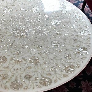 May include: A round white marble table with an intricate floral inlay design. The inlaid pattern features silver-colored floral and leaf motifs, creating a detailed and elegant surface. The table's edge is smooth and rounded.