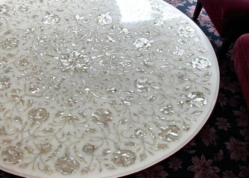 May include: A round white marble table with an intricate floral inlay design. The inlaid pattern features silver-colored floral and leaf motifs, creating a detailed and elegant surface. The table's edge is smooth and rounded.