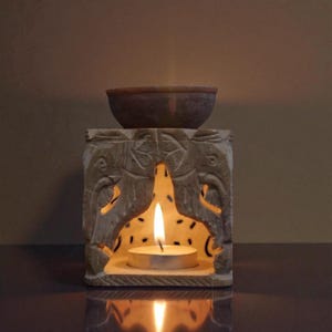 Puede incluir: Un difusor de aceites esenciales de piedra tallada con diseños de elefantes. Una pequeña taza se encuentra en la parte superior, y una vela de té encendida brilla en el interior. El difusor es de color gris claro, y el fondo es marrón cálido.