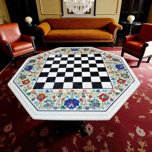 May include: An octagonal white marble coffee table with a black and white chessboard in the centre. The table is decorated with colourful floral patterns around the chessboard. The table is supported by black legs.