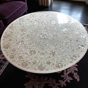 May include: A round white marble coffee table with an intricate floral inlay design. The table's surface is adorned with detailed silver-toned floral patterns. The table is set on a dark rug with a purple floral border, and a purple sofa is visible in the background.
