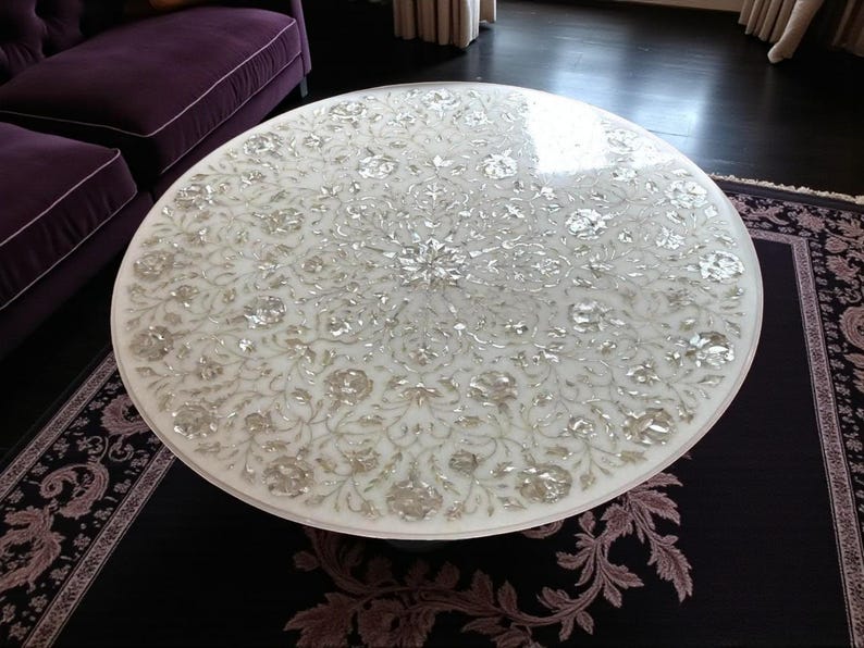 May include: A round white marble coffee table with an intricate floral inlay design. The table's surface is adorned with detailed silver-toned floral patterns. The table is set on a dark rug with a purple floral border, and a purple sofa is visible in the background.