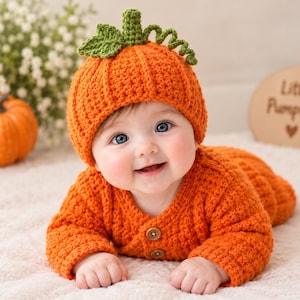 May include: A bright orange crochet baby outfit consisting of a long-sleeved top with two brown buttons and a matching hat featuring a green stem, a leaf, and a curly vine. The textured yarn garment is designed with a pumpkin-inspired aesthetic.