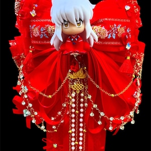 May include: A red figurine with white hair and yellow eyes, adorned with a red dress and elaborate gold and pearl jewelry. The figurine is set against a backdrop of red fabric with gold embroidery and decorative tassels.