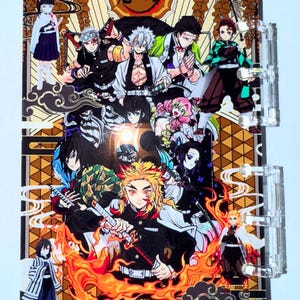 Op de afbeelding: Een doorzichtige plastic binder met een kleurrijke illustratie van meerdere anime-personages. Het kunstwerk bevat personages met zwaarden, vlammen en diverse outfits. De achtergrond heeft een goud-bruin geometrisch patroon met Japanse tekst.