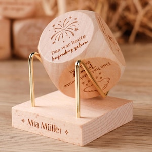 Puede incluir: Un cubo de madera de color claro con iconos grabados y texto en alemán, incluyendo 'Das war heute besonders schön', sobre un soporte de metal dorado unido a una base cuadrada de madera grabada con el nombre 'Mia Müller'.