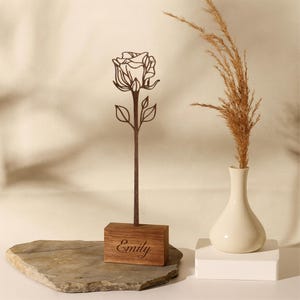 以下が含まれることがあります： ベースに「Emily」と刻まれた木製のバラの彫刻。バラは茶色で、詳細なアウトラインがあります。右側には、ドライグラスが入った白い花瓶があります。彫刻は石の上にあります。