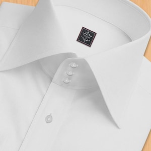 May include: A close-up of a white dress shirt with a high collar and three small buttons near the collar. The shirt features a small label with the word "PANACHE". The shirt is made of a smooth, likely cotton, fabric.