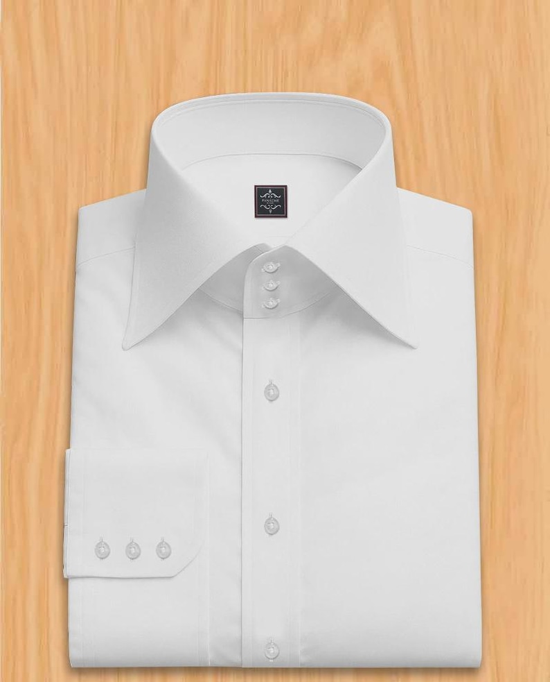 May include: A pristine white dress shirt, featuring a distinctive high collar and French cuffs. The shirt is neatly folded, highlighting the buttoned placket and cuff details. The background is a light wood grain.