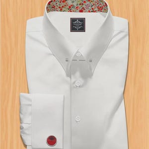 Puede incluir: Una camisa blanca con forro de cuello estampado floral y detalles en los puños. La camisa presenta un cuello con alfiler, cierre de botones y gemelo rojo. La camisa está cuidadosamente doblada sobre una superficie de madera clara. La etiqueta dice "PANACHE".