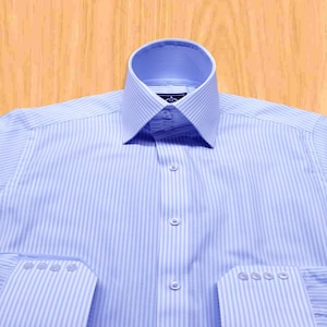 Puede incluir: Camisa azul claro con rayas blancas verticales. La camisa tiene cuello italiano, cierre de botones y puños abotonados. Los puños tienen cuatro botones cada uno. Fabricada en tejido ligero.
