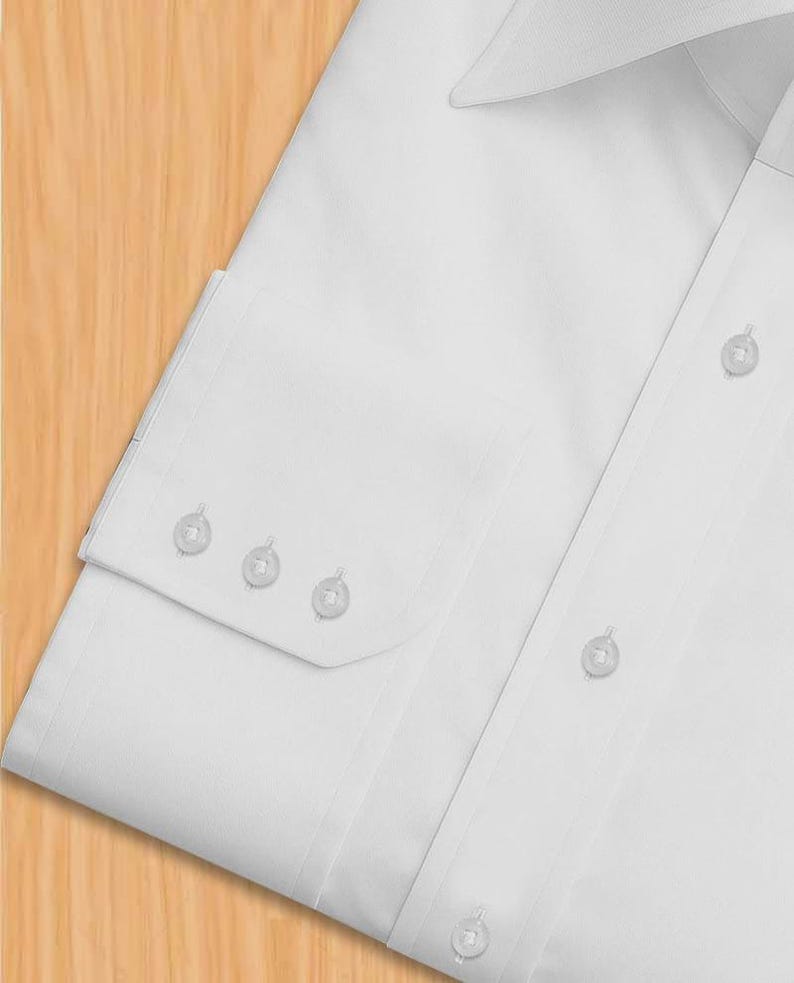 May include: A close-up view of a white dress shirt. The shirt has a classic collar, a button-down front, and a buttoned cuff with three buttons. The fabric looks smooth and crease-free, indicating a high-quality material.
