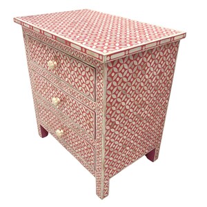 May include: A red and white patterned nightstand with three drawers. The nightstand features a geometric design with white floral shapes on a red background. The drawer pulls are white, and the legs are a matching red color. The overall dimensions are approximately 24 inches wide, 16 inches deep, and 30 inches tall.
