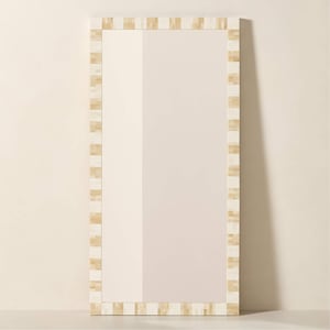 Puede incluir: Un espejo rectangular con un marco color crema. El marco está decorado con un patrón repetitivo de piezas rectangulares en tonos crema y beige. El espejo está apoyado contra una pared de color claro.