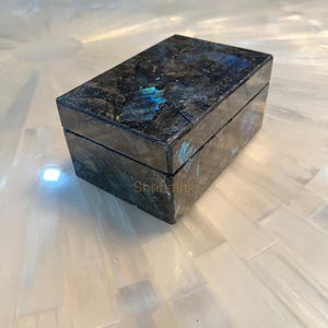 以下が含まれることがあります： 暗く虹色のラブラドライト石で作られた長方形のジュエリーボックス。ボックスはヒンジ付きの蓋と研磨された仕上げで、青と緑の閃光があります。ボックスは白い表面にあります。「Shrinathji」というテキストが表示されます。