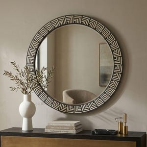 May include: A round wall mirror featuring a dark frame with a repeating white geometric chevron pattern. The mirror reflects a room interior, including a chair. The design consists of concentric angular lines creating a structured, repetitive motif.