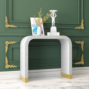 May include: A white and silver console table with a curved design and gold-coloured accents. The table is decorated with a white statue, a framed picture, and a vase with yellow flowers. The table is 122 cm long, 41 cm wide, and 76 cm high.