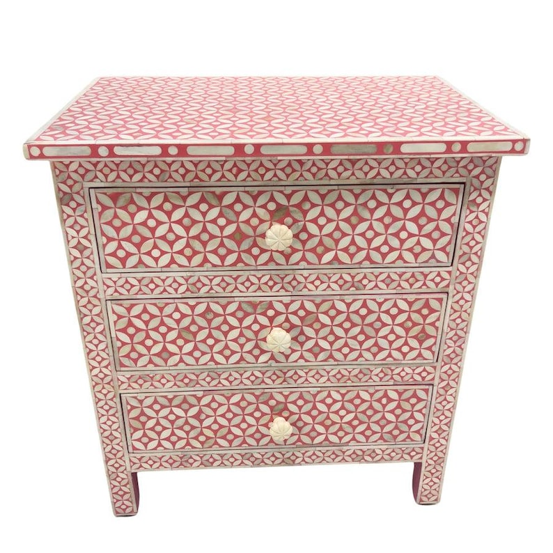 May include: A red and white patterned three-drawer chest. The chest features a geometric design on the top and drawer fronts, with white floral knobs. The chest is a decorative piece of furniture.
