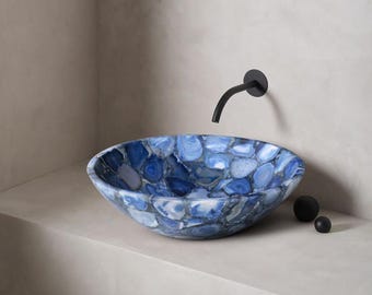 May include: Oval-shaped blue and white stone sink with a black faucet. The sink is made of polished stones in shades of blue and white. Two small black spheres are on the counter next to the sink. The sink is mounted on a light gray surface.
