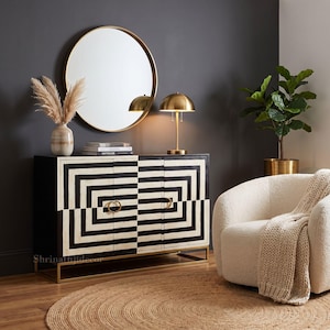 May include: A rectangular sideboard featuring a black and white geometric pattern on its three doors, accented with gold ring pulls and a gold metal base. The piece is topped with a stack of books, a vase with dried grass, and a gold dome lamp.
