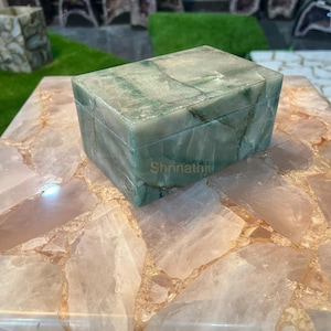 Könnte beinhalten: Eine rechteckige, grün getönte Steinbox mit Deckel, die auf einer hellrosa und weißen Oberfläche ruht. Die Box weist sichtbare Maserungen auf und das Wort "Shrinathji" ist auf der Seite zu sehen. Der Hintergrund umfasst weitere Steinstücke und grünen Rasen.