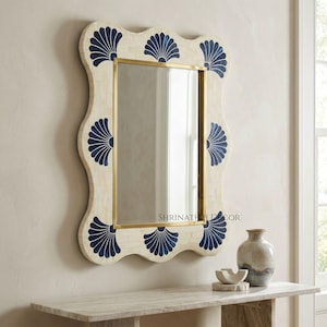 May include: A rectangular wall mirror with a cream-colored frame, adorned with navy blue fan-shaped designs. The mirror has a gold-colored trim and a wavy edge. The mirror is hanging on a wall.