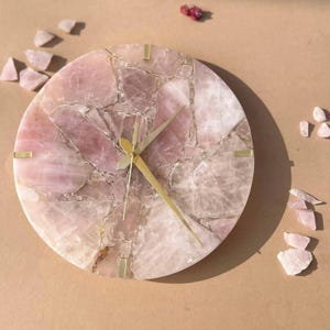 Puede incluir: Un reloj redondo con una esfera de cuarzo rosa. El reloj tiene manecillas y marcadores dorados. El cuarzo rosa es de color rosa claro con vetas rosas y blancas más oscuras. Pequeños trozos de cuarzo rosa están esparcidos alrededor del reloj.
