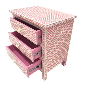 May include: A pink three-drawer nightstand with a geometric pattern. The drawers are open, revealing a pink interior. The knobs are cream-colored. The nightstand is made of wood and has a decorative design.