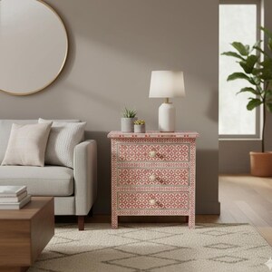 May include: A three-drawer nightstand with a pink and white geometric pattern. The nightstand has a white lamp, two small potted plants, and a white lamp. The room also includes a gray sofa, a round mirror, and a patterned rug.