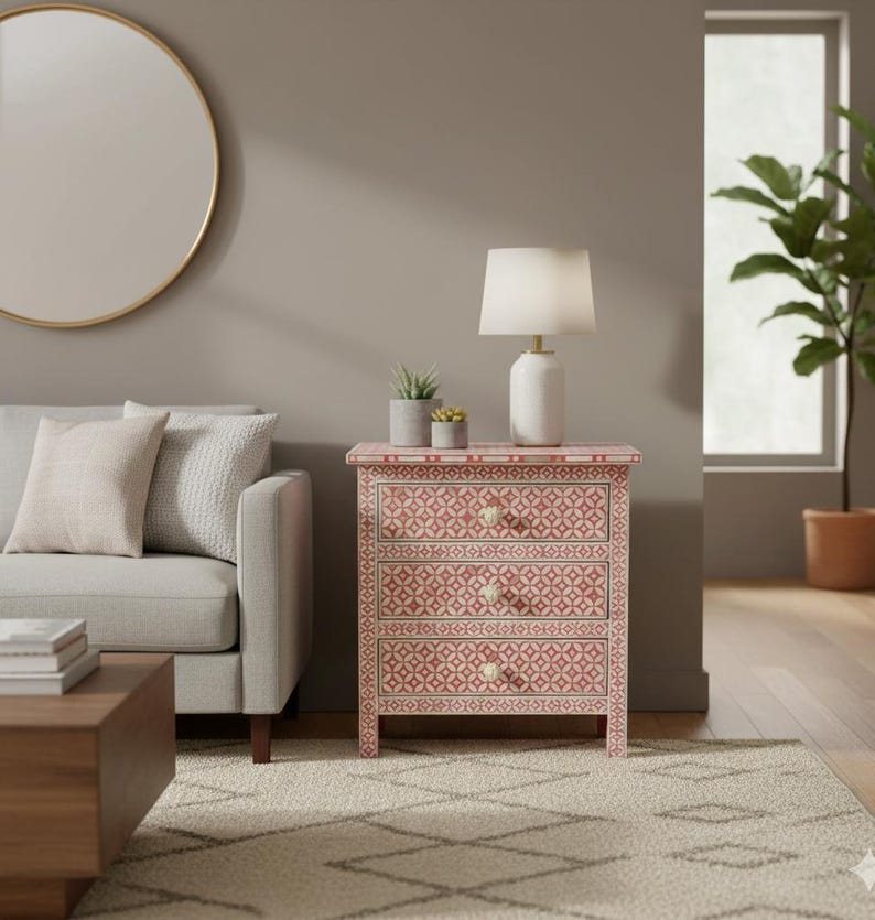 May include: A three-drawer nightstand with a pink and white geometric pattern. The nightstand has a white lamp, two small potted plants, and a white lamp. The room also includes a gray sofa, a round mirror, and a patterned rug.