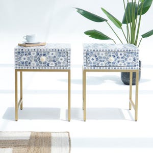 May include: Two matching side tables with a mosaic tile design in shades of blue and white. Each table has a single drawer with a gold-coloured knob and gold-coloured metal legs. A cup and saucer sit on one table.