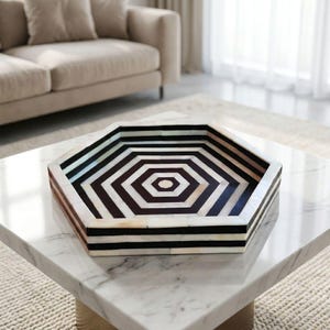 May include: A hexagonal tray featuring a geometric pattern of alternating black and cream-colored segments. The tray has raised sides with a repeating striped design that follows the hexagonal shape, creating a concentric visual effect.