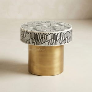 May include: A round side table featuring a top and side panel with a black and white geometric cube pattern. The base is a solid, cylindrical gold-coloured metal pedestal. The text Shrinathjidecor is visible on the side of the table base.
